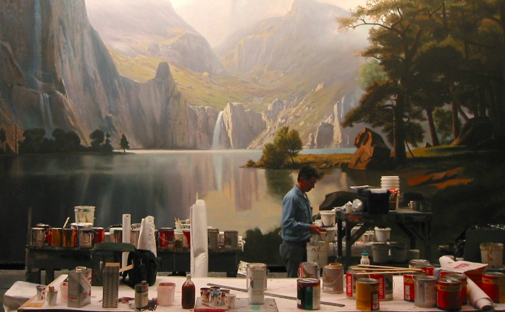 A scenic painter prepares his paint and tools in front of an epic landscape backdrop showcasing the Scenic Paint Department Roles